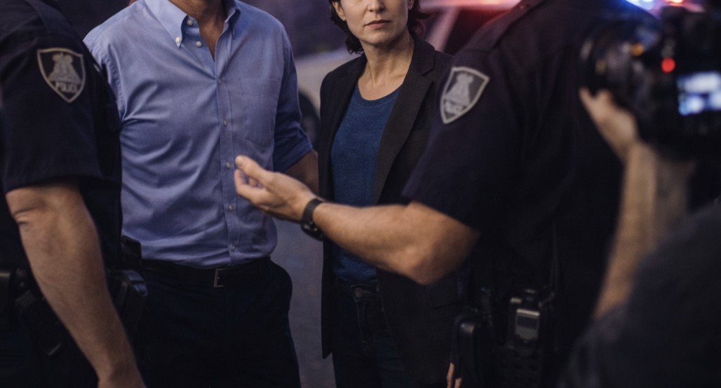 Police officers speak with a man and a woman during a nighttime interaction, with flashing patrol lights visible in the background.